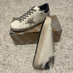 Vita sneakers Golden goose sneakers - Snygga vita sneakers från Golden Goose med en grå stjärna på sidan och mörkblå detaljer vid hälen. Skorna har grå snörning och en vintage-look på sulan. Perfekta för en stilren och avslappnad stil. Medela vid intresse 🤗