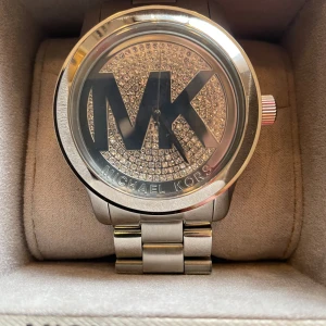 Silverfärgad klocka från Michael Kors - Snygg och elegant klocka från Michael Kors med ett stort MK-logotyp på urtavlan. Klockan har en glittrig bakgrund och ett robust armband i rostfritt stål. Perfekt för att ge en touch av glamour till din stil.