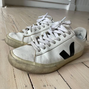 Vita Veja sneakers med svart detalj - Säljer ett par vita Veja sneakers med svart detalj på sidan. Skorna har snörning och en klassisk design med rund tå. Perfekta för en avslappnad stil.