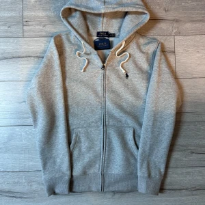 Grå hoodie från Polo Ralph Lauren - Säljer en stilren grå hoodie från Polo Ralph Lauren med dragkedja och justerbar huva. Tröjan har ribbade muddar och en liten broderad logga på bröstet. Perfekt för en avslappnad look.