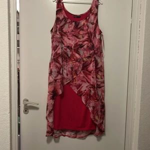 Rosa mönstrad klänning från Bodyflirt - Säljer en snygg rosa klänning från Bodyflirt med ett abstrakt mönster i olika nyanser av rosa och rött. Klänningen är ärmlös och har en asymmetrisk design med ett lager över kjolen. Perfekt för en färgglad look! Har en liten fläck nertill (se bild två).