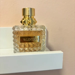 Parfym från Valentino - Elegant parfymflaska från Valentino med en lyxig design. Flaskan är fyrkantig och har ett mönster av små pyramider i glaset. Den har en gyllene etikett och ett matchande gyllene lock med nitar. Perfekt för den som älskar exklusiva dofter.