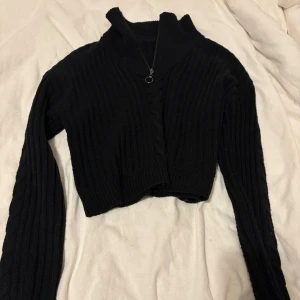 Svart ribbad croppad tröja med dragkedja - Säljer en svart ribbad croppad tröja med dragkedja och hög krage. Perfekt för en stilren look. Tröjan har långa ärmar och är gjord i ett mjukt material.