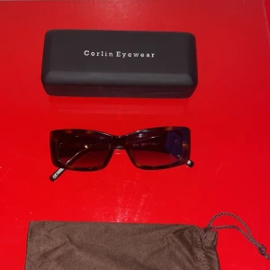 Bruna solglasögon från Corlin Eyewear - Snygga rektangulära solglasögon från Corlin Eyewear i en stilren brun färg. Perfekta för att ge en cool touch till din outfit. Kommer med ett praktiskt fodral för säker förvaring. Ordinarie pris - 1 400 SEK