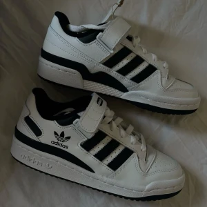 Vita Adidas forum  - Snygga vita sneakers från Adidas med svarta detaljer och klassiska tre ränder på sidan. Skorna har snörning och kardborreband för extra stöd. Perfekta för en sportig look. 