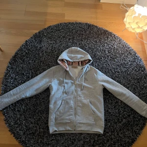 Burrberry Hoodie - Säljer en stilren grå hoodie från Burberry med klassiskt rutigt foder i huvan. Tröjan har dragkedja och fickor framtill, perfekt för en avslappnad look. Tillverkad i bomull med en liten del elastan för extra komfort. Perfekt till våren och sommaren. Priset går att förhandla över och om du har en fråga? Fråga gärna på då :)