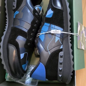 Valentino Garavani sneakers i blått och svart - Snygga Valentino Garavani sneakers med ett coolt blått och svart camouflagemönster. Skorna har svarta snören och en robust sula med nitar på hälen. Perfekta för en stilren look med en twist. Ej använda samt helt nya. Pris kan diskuteras vid snabb affär.