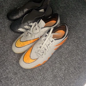 Fotbollsskor från Nike och Adidas - Två par fotbollsskor till salu. Ett par Nike i grått med orange detaljer och snörning. Det andra paret är Adidas Predator i svart med vita detaljer och utan snörning. Svarta storlek 35, dom gråa storlek 38