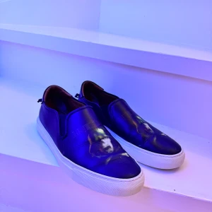 Svarta slip-on sneakers från Givenchy - Snygga svarta slip-on sneakers från Givenchy med vit sula och subtilt mönster på ovansidan. Skorna har en elegant design med Givenchy-logga på hälen och sulan. Perfekta för en stilren look.