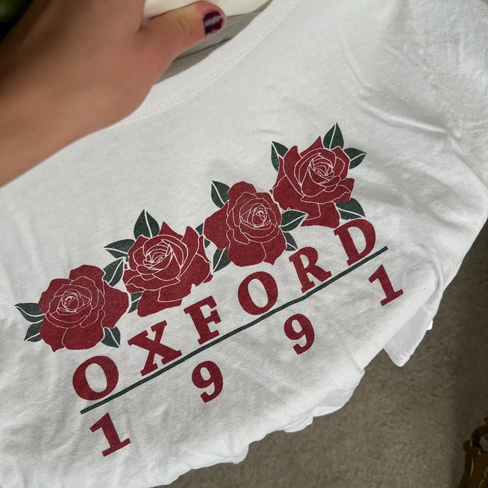 Säljer en vit t-shirt med tryck av röda rosor och texten 'Oxford 1991'. T-shirten har en croppad stil och korta ärmar, perfekt för en avslappnad look. Passar mellan xs-m, bra skick men lite skrynklig . T-paidat.