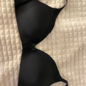 Svart bh med spetsdetaljer - Snygg svart bh med spetsdetaljer och en liten rosett framtill. Den har justerbara axelband och en klassisk knäppning bak. Bh-n har aldrig används för jag gillar inte modellen. 