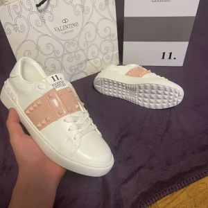 Valentino Rockstud skor - Snygga vita Valentino Rockstud sneakers med rosa detaljer och ikoniska nitar. Skorna har en stilren design med snörning och en bekväm passform. Perfekta för att ge din outfit en lyxig touch.