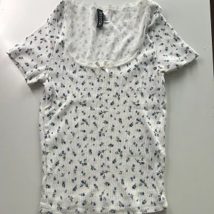 Vit blommig topp från H&M - Söt vit topp med små blå blommor från H&M. Den har korta ärmar och en fin spetskant runt halsringningen samt en liten rosett framtill. Perfekt för en somrig look! 100kr +frakt pris kan diskuteras