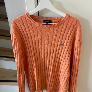 Orange stickad tröja från GANT - Säljer en  orange stickad tröja från GANT med kabelstickat mönster. Tröjan har en rund halsringning och ett litet broderat emblem på bröstet. 