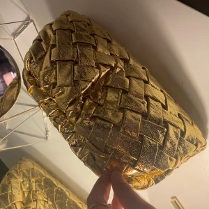 Guld kuvertväska i flätat mönster - Snygg guld kuvertväska med ett elegant flätat mönster. Perfekt för att ge en lyxig touch till din outfit. Väskan är kompakt och lätt att bära med sig.