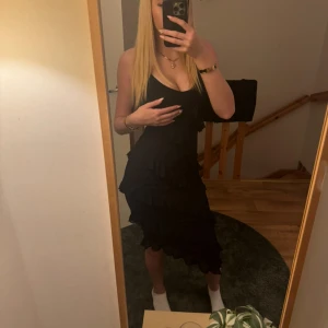 Svart klänning med volanger - Snygg svart klänning med volanger och smala axelband. Perfekt för en kväll ute eller en speciell tillställning. Klänningen har en elegant och stilren design som framhäver figuren.