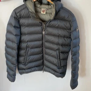 Grå dunjacka från Colmar - Säljer en stilren grå dunjacka från Colmar. Jackan har en praktisk huva och dragkedja framtill. Perfekt för kyligare dagar med sin quiltade design som håller värmen. Två fickor med dragkedja på framsidan för extra funktionalitet.❣️