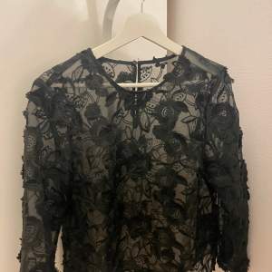 Elegant svart blus i genomskinlig spets med vackert blommönster. Blusen har långa ärmar och en rund halsringning, perfekt för en stilfull look.