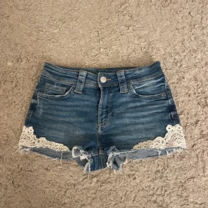 Jeansshorts med spetsdetaljer - Snygga jeansshorts med fransiga kanter och vita spetsdetaljer längs benen. Perfekta för en avslappnad sommarlook. De har en klassisk femficksdesign och knappgylf.