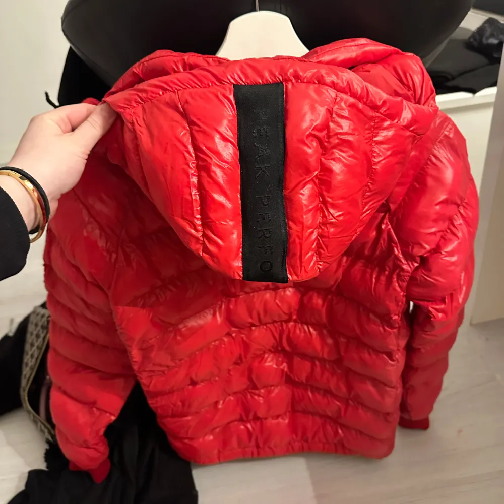 Säljer en snygg röd dunjacka från Peak Performance. Jackan har en dragkedja framtill och en huva för extra värme. Perfekt för kyliga dagar med sin stilrena design och praktiska fickor. Märkesdetaljer på ärmen ger en sportig touch.. Takit.