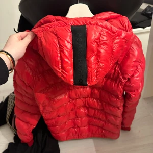 Röd dunjacka från Peak Performance - Säljer en snygg röd dunjacka från Peak Performance. Jackan har en dragkedja framtill och en huva för extra värme. Perfekt för kyliga dagar med sin stilrena design och praktiska fickor. Märkesdetaljer på ärmen ger en sportig touch.