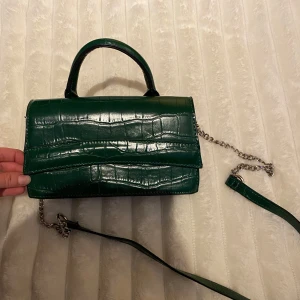 Grön axelväska med kedja - Snygg mörk grön axelväska med krokodilpräglat mönster och silverfärgad kedja. Väskan har ett handtag och en justerbar axelrem. Perfekt för att ge en färgklick till din outfit!