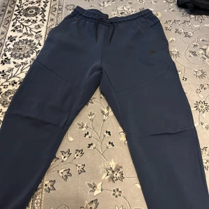 Mörkblå Nike tech fleece byxor  - Säljer ett par mörkblåa Nike tech fleece bysor med elastisk midja och dragsko. Byxorna har en dragkedjeficka på sidan för extra förvaring. Perfekta för en avslappnad stil. Knappt använda och inga skador på dem alls. Priset kan diskuteras!