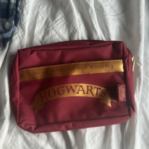 Röd necessär med Hogwarts-tryck - Snygg röd necessär med Hogwarts-tryck i guld. Perfekt för alla Harry Potter-fans som vill ha en praktisk och stilren förvaring. Necessären har dragkedja och är lätt att ta med sig.