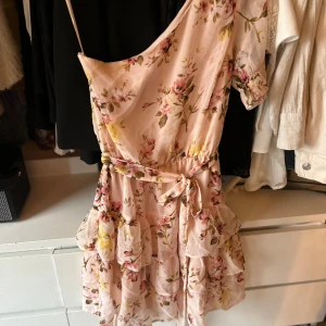 Blommig klänning från NLY Trend - Söt blommig klänning från NLY Trend i ljusrosa med volanger och knytband i runt magen. Klänningen har en offshoulder-design och kort längd.