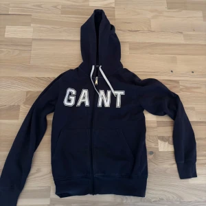Mörkblå hoodie från GANT - Säljer en snygg mörkblå hoodie från GANT med dragkedja och stor logga på bröstet. Den har långa ärmar och en praktisk huva med snören. Perfekt för en avslappnad stil. Obs ett av snörrena är lite sönder