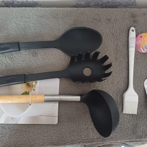 Köksredskap set med slevar och osthyvel - Ett set med köksredskap inklusive två svarta slevar, en pastaslev, en soppslev med trähandtag, en vit bakpensel och en rostfri osthyvel. Perfekt för matlagning och servering i köket.