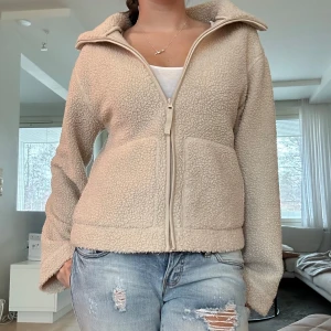 Beige teddyjacka - Mysig beige teddyjacka med dragkedja framtill och två fickor. Perfekt för kyliga dagar när du vill hålla dig varm och bekväm. Jackan har en avslappnad passform och långa ärmar.