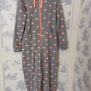 Grå onesie med hjärtan - Mysig grå onesie med rosa hjärtan över hela plagget. Den har en dragkedja framtill och en huva med snören. Perfekt för en avslappnad dag hemma.