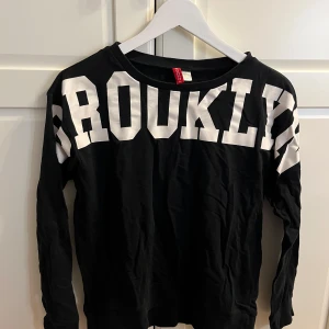 Svart långärmad tröja från H&M Divided - Säljer en svart långärmad tröja från H&M Divided med texten 'BROOKLYN' i stora vita bokstäver över bröstet. Perfekt för en avslappnad stil. Tröjan har en rund halsringning och är tillverkad i ett mjukt material.