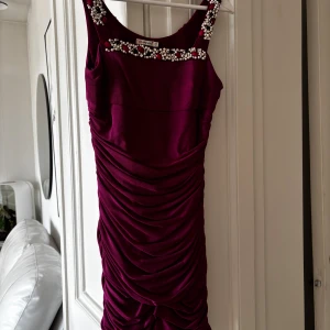 Vinröd klänning från Chi-Chi - Snygg vinröd klänning från Chi-Chi med pärldetaljer vid halsringningen. Klänningen har en draperad design som ger en elegant look. Perfekt för speciella tillfällen.