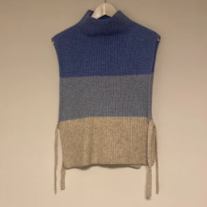 Blå och beige stickad väst - Säljer en stilren stickad väst i blått och beige. Västen har en hög krage och knytband på sidorna för en justerbar passform. Perfekt för lager-på-lager under kyligare dagar.