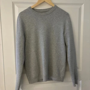 Grå stickad tröja - Säljer en stilren grå stickad tröja med rund halsringning. Tröjan har långa ärmar och en ribbad kant vid halsen. Perfekt för kyligare dagar.