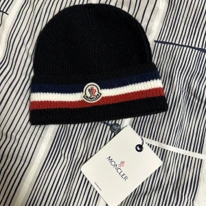 Svart mössa från Moncler - Snygg svart mössa från Moncler med ribbad design och ränder i blått, vitt och rött. Den har en broderad Moncler-logga framtill. Perfekt för att hålla sig varm med stil.