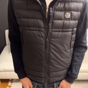 Svart dunväst från Stone Island - Snygg svart dunväst från Stone Island med dragkedja framtill. Västen är tillverkad i bio-baserad ripstop nylon och har en stilren design med märkets logga på bröstet. Perfekt för att hålla värmen med stil.