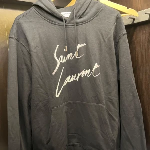 Sällsynt saint laurent hoodie  - Säljer en stilren svart hoodie från Saint Laurent med deras ikoniska logga i vitt på framsidan. Tröjan har en klassisk design med huva och dragsko samt en praktisk magficka. Perfekt för en avslappnad look. Säljer pågrund av att den är för stor. Den är i nyskick. 