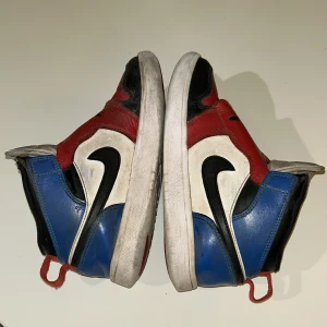 Sky Jordan sneakers i rött, blått och svart - Säljer ett par Sky Jordan sneakers i en cool färgkombination av rött, blått och svart. Skorna har en ikonisk design med en stor logotyp på sidan och perforerade detaljer på tån. Perfekta för dig som vill ha en sportig och stilren look. En av skon är lite synligt sönder men syns inte så mycket!
