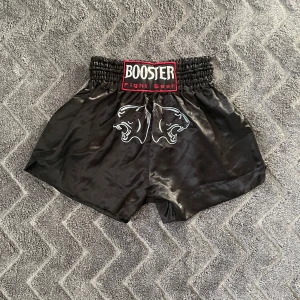  Booster kickboxing shorts  - Snygga svarta träningsshorts från Booster Fight Gear med elastisk midja och coolt tryck av två pantrar framtill. Perfekta för intensiva träningspass eller kampsport.