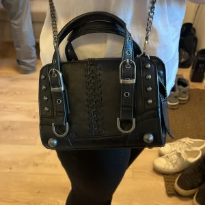 Svart axelväska med kedjedetaljer - Snygg svart axelväska i skinn med kedjedetaljer och nitar. Väskan har ett snörningsmönster på framsidan och två handtag och ett längre band 🌸 