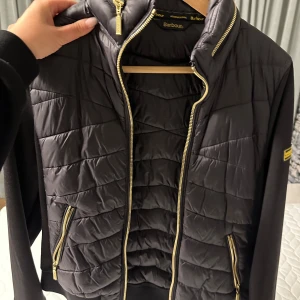 Svart quiltad jacka från Barbour - Säljer en snygg svart quiltad jacka från Barbour International. Jackan har en dragkedja i guld och ribbade muddar. Perfekt för en stilren look med en touch av motorcykelkänsla.
