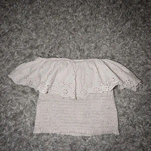 Beige offshoulder topp - Säljer en söt beige offshoulder topp med volangdetaljer och broderade mönster. Toppen är croppad och har en smockad design som ger en tight passform. Perfekt för sommarens varma dagar! Aldrig använd! Köpt i en marknad i Spanien! SITTER EXAKT SOM ANDRA BILDEN 