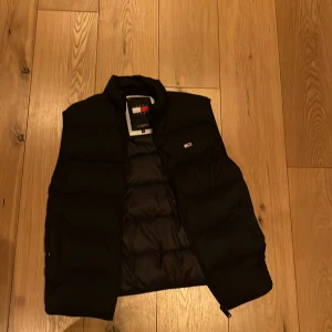 Svart dunväst från Tommy Hilfiger - Snygg svart dunväst från Tommy Hilfiger med en stilren design. Västen har en diskret logotyp på bröstet och en hög krage för extra värme. Perfekt för lager-på-lager under kyliga dagar.