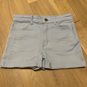 Ljusblå shorts från H&M - Säljer ett par ljusblå shorts från H&M. De har en klassisk design med uppvikta ben och knappstängning fram. Shortsen har både fram- och bakfickor, vilket gör dem praktiska och stilrena.