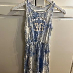 Blå och vit klänning från H&M - Säljer en cool blå och vit klänning från H&M med texten 'California 36' på framsidan. Klänningen har en avslappnad passform och ärmlös design, perfekt för varma dagar. Den har ett elastiskt band i midjan för en bekväm passform.