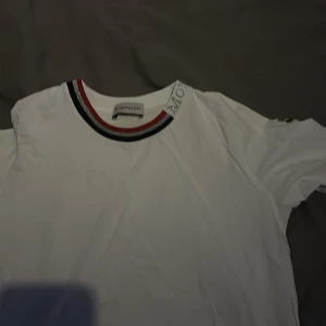 Vit långärmad t-shirt från Moncler - Säljer en stilren vit långärmad tröja från Moncler. Den har en rund halsringning med ränder i blått, rött och svart samt Moncler-logga på kragen. Perfekt för en avslappnad och trendig look. Liten i storlek, pris kan diskuters. STORLEK 152