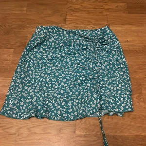 Blommig kjol från Shein - Snygg blommig kjol från Shein i en härlig grön/blå färg med vita blommor. Jag skulle säga att den är turkos. Kjolen har en knytbar detalj framtill som ger en fin rynkad effekt. Man kan även dra i snörret så den bli kortare🤗Perfekt för sommarens alla tillfällen!
